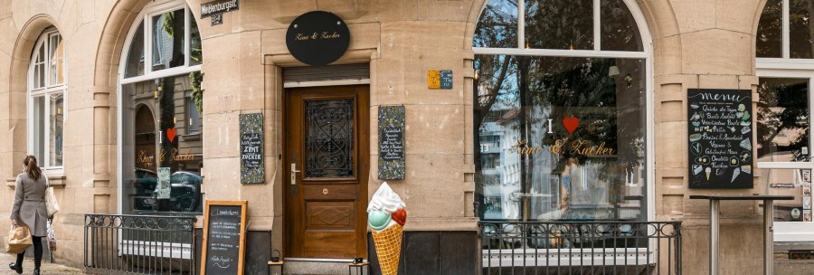 Ein Caf&eacute; namens 'Zimt & Zucker' an einer Stra&szlig;enecke mit gro&szlig;en Fenstern, einem Eist&uuml;tenmodell und einer Frau, die vorbeigeht., &copy; SMG, Sarah Schmid