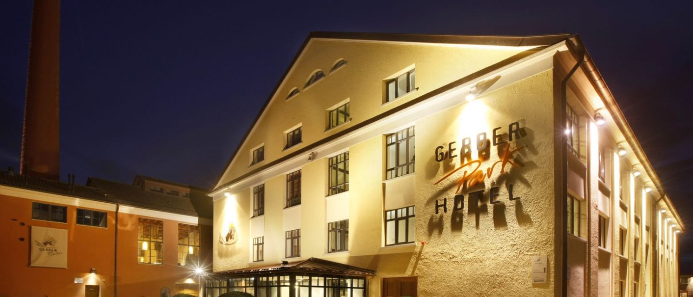 Das Hotel Gerber-Park bei Nacht, beleuchtet mit gelber Fassade und markantem Schriftzug. Im Hintergrund ein hoher Schornstein., © TOMAS Das Hotel Gerber-Park bei Nacht, beleuchtet mit gelber Fassade und markantem Schriftzug. Im Hintergrund ein hoher Schornstein., © TOMAS