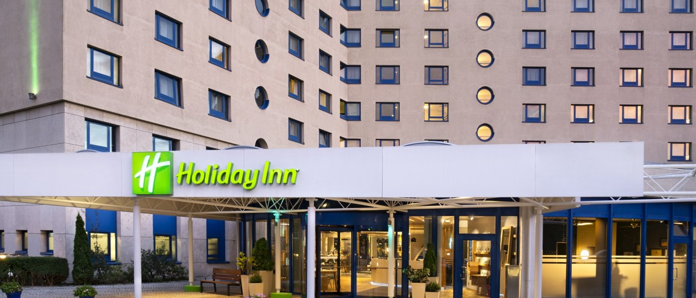 Exterior view of a Holiday Inn hotel with illuminated entrance, round and rectangular windows. Paved forecourt with plants., &copy; Holiday Inn Stuttgart