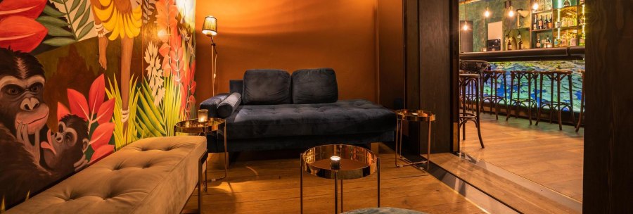 Cosy lounge with blue couch, colorful mural with monkeys and plants. A bar with illuminated shelves and stools in the background., © Holzmaler, Stuttgart