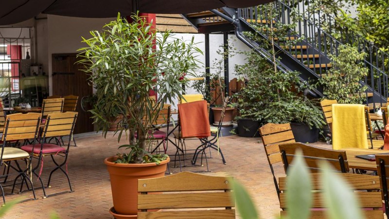 Gem&uuml;tlicher Caf&eacute;-Au&szlig;enbereich mit Holztischen und -st&uuml;hlen, umgeben von Pflanzen. Bunte Decken auf den St&uuml;hlen sorgen f&uuml;r eine einladende Atmosph&auml;re., &copy; SMG, Sarah Schmid
