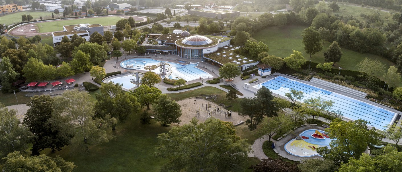 Luftaufnahme des Wunnebads in Winnenden: Schwimmbecken, Rutschen und grüne Umgebung. Sportplatz im Hintergrund., © Stadtwerke Winnenden GmbH