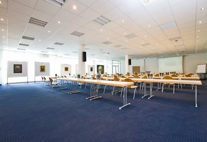 A bright conference room with long tables and chairs, blue carpet, pictures on the walls, and a projection screen at the front., &copy; bonjour Hotelbetriebs GmbH