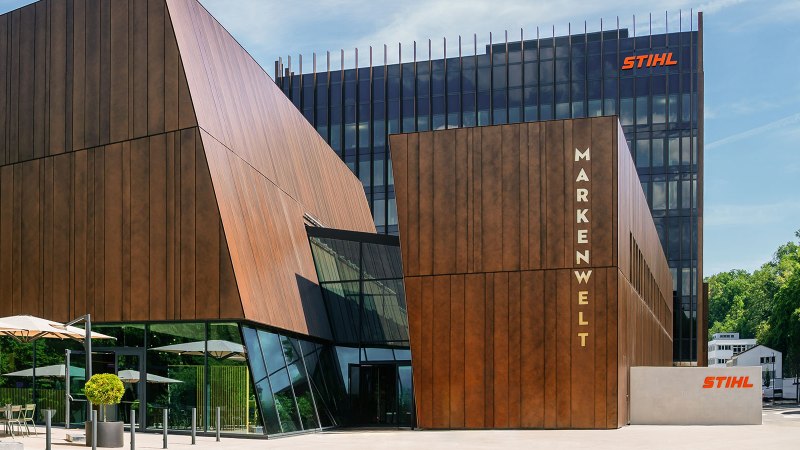 Modern STIHL brand world building with wooden cladding and glass fronts, surrounded by trees. Sunshades and seating in the foreground., © STIHL Markenwelt Modern STIHL brand world building with wooden cladding and glass fronts, surrounded by trees. Sunshades and seating in the foreground., © STIHL Markenwelt