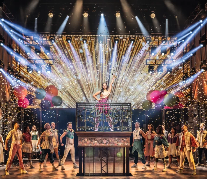 A woman is standing on a platform in a musical, surrounded by dancers and confetti. Colorful lights and balloons decorate the stage., © Johan Persson / Stage Entertainment A woman is standing on a platform in a musical, surrounded by dancers and confetti. Colorful lights and balloons decorate the stage., © Johan Persson / Stage Entertainment