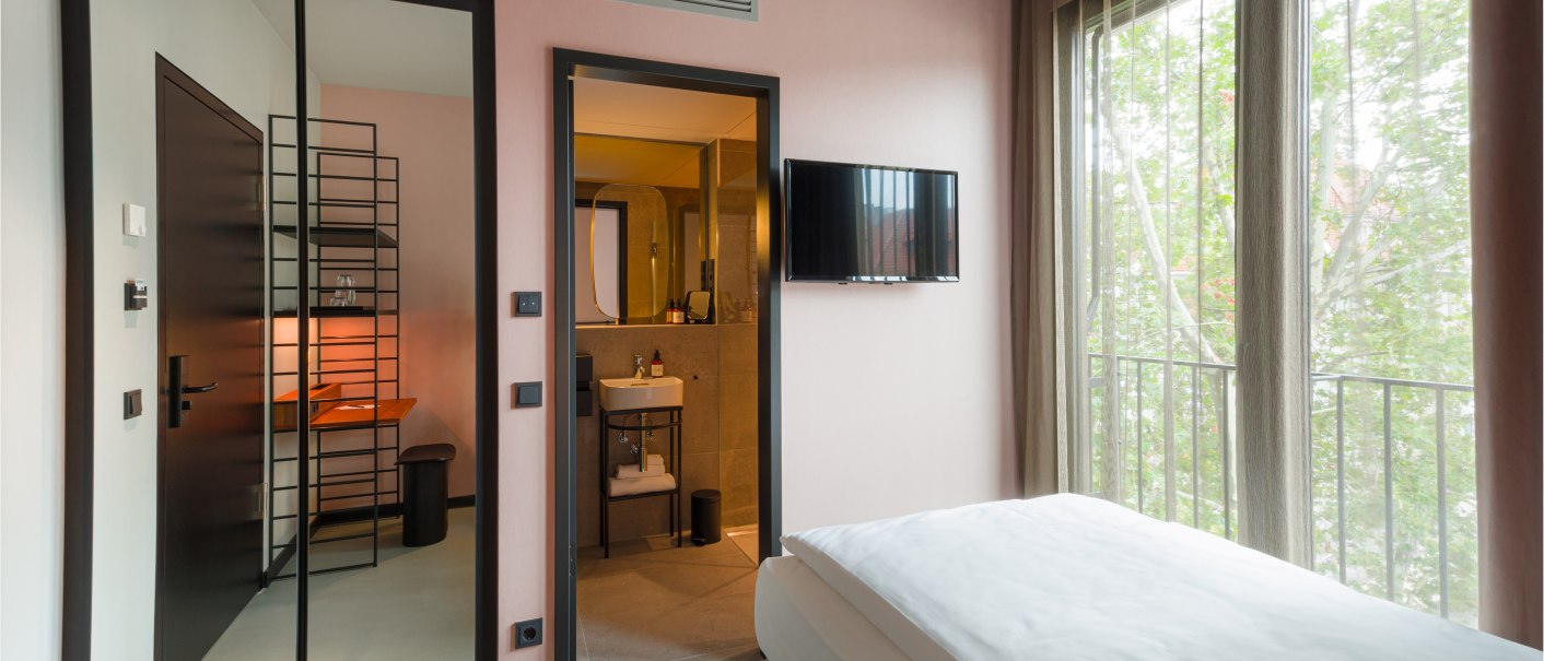 Modernes Zimmer mit Bett, Fernseher an der Wand, großem Fenster mit Vorhängen, Spiegel und offenem Badezimmer mit Waschbecken., © EmiLu Hotel GmbH
