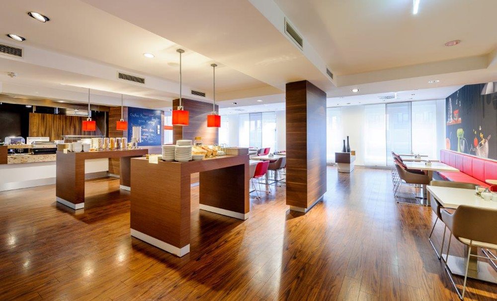 Moderner Frühstücksbereich mit Holzboden, Tischen, roten Lampen und Sitzgelegenheiten. Helle, einladende Atmosphäre mit Buffetbereich., © Ibis Stuttgart Airport Messe