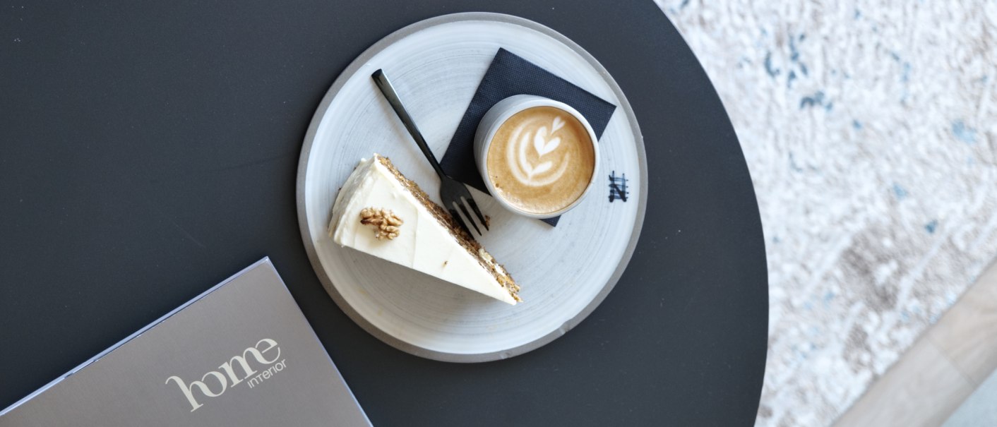 Latte mit Herz-Latte-Art und ein Stück Kuchen auf einem Teller auf schwarzem Tisch, daneben eine Karte mit „home interior“., © Taraba Latte mit Herz-Latte-Art und ein Stück Kuchen auf einem Teller auf schwarzem Tisch, daneben eine Karte mit „home interior“., © Taraba