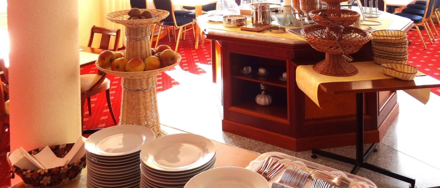 Ein Frühstücksraum mit Buffet, gestapelten Tellern, Besteck und Obstkörben auf einem Tisch. Im Hintergrund sind Tische und Stühle zu sehen., © Hotel Arcis