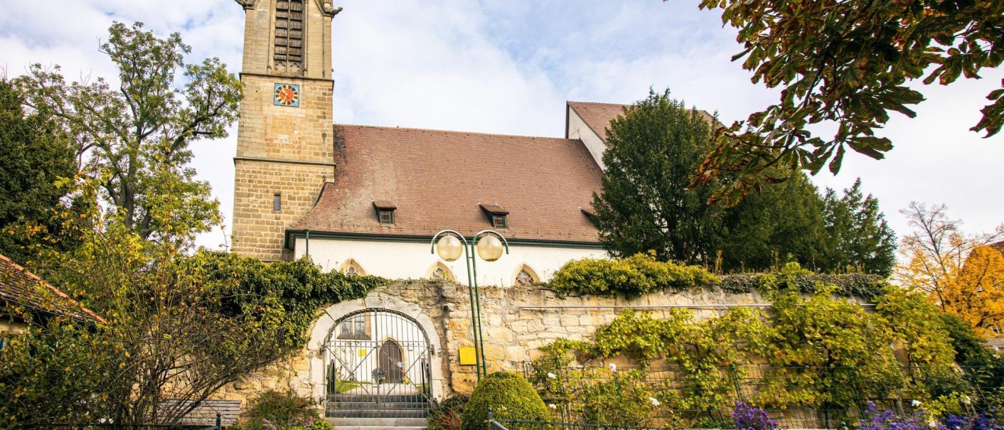 A historic church in Leinfelden-Echterdingen, surrounded by lush greenery and autumnal trees. A gate leads to the entrance of the church., © Stuttgart-Marketing GmbH, Sarah Schmid