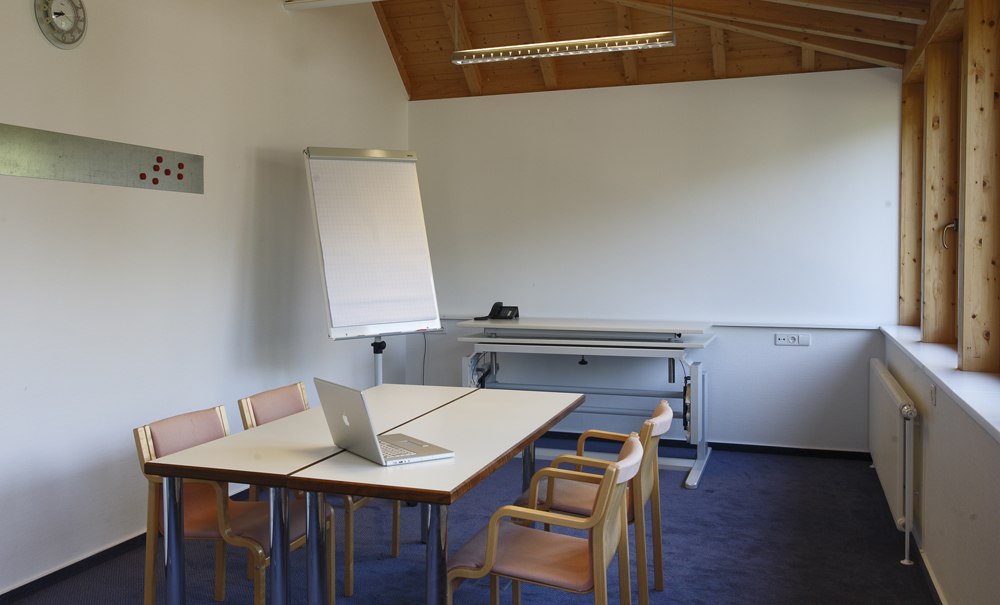 Ein Seminarraum mit Holzdach, Tisch, Stühlen, Flipchart und Laptop. Große Fenster lassen viel Licht herein. Eine Uhr hängt an der Wand., © Badhotel Restaurant Stauferland