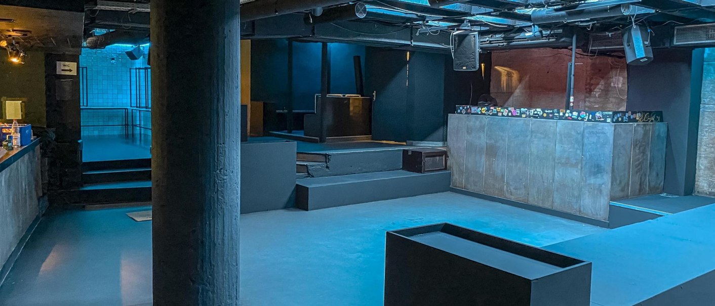 Ein leerer Nachtclub mit blauer Beleuchtung, DJ-Pult und Bar. Der Raum wirkt modern und industriell., © Schräglage, Stuttgart