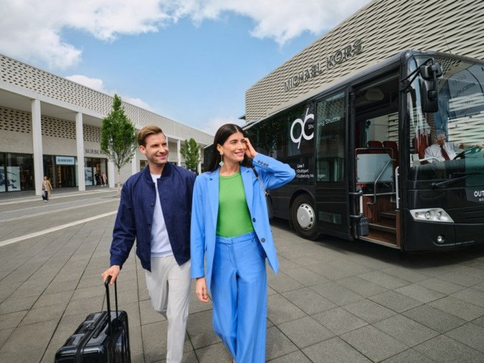 Ein Paar mit Koffern läuft vor einem Bus in der Outletcity Metzingen. Im Hintergrund sind Geschäfte und ein Michael Kors Laden zu sehen., © Schlienz-Tours GmbH & Co.KG Ein Paar mit Koffern läuft vor einem Bus in der Outletcity Metzingen. Im Hintergrund sind Geschäfte und ein Michael Kors Laden zu sehen., © Schlienz-Tours GmbH & Co.KG
