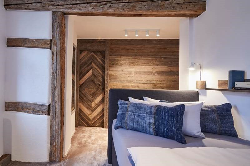 Schlafzimmer mit Holzbalken, modernem Bett, blauen Kissen und Holzverkleidung. Warmes Licht sorgt f&uuml;r eine gem&uuml;tliche Atmosph&auml;re., &copy; Hotel Restaurant Bachofer