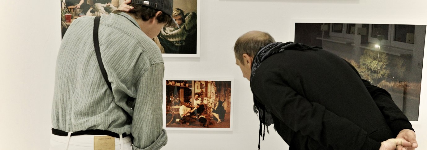 Zwei Personen betrachten aufmerksam Fotos an einer wei&szlig;en Galeriewand. Die Bilder zeigen verschiedene Szenen, darunter Menschen und eine Schneelandschaft., &copy; Palermo Galerie