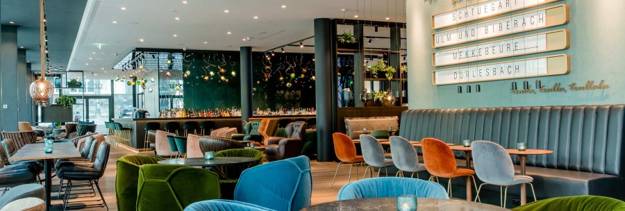 Modernes Restaurant mit bunten Sesseln, Tischen und stilvoller Beleuchtung. Eine Tafel an der Wand zeigt St&auml;dtenamen. Elegante und gem&uuml;tliche Atmosph&auml;re., &copy; Motel One - Stuttgart Hauptbahnhof