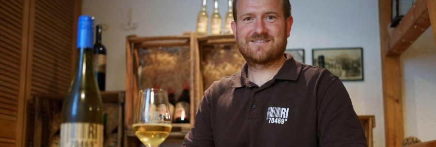 A man in a wine cellar smiles at the camera and holds a glass of white wine. Wine bottles can be seen in the background., &copy; 70469R!