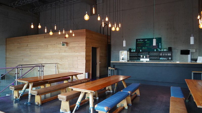 Ein modernes Café mit Holztischen und Bänken, stilvollen Hängelampen und einer Bar im Hintergrund. Die Atmosphäre ist einladend und gemütlich., © Württembergische Staatstheater Stuttgart Ein modernes Café mit Holztischen und Bänken, stilvollen Hängelampen und einer Bar im Hintergrund. Die Atmosphäre ist einladend und gemütlich., © Württembergische Staatstheater Stuttgart