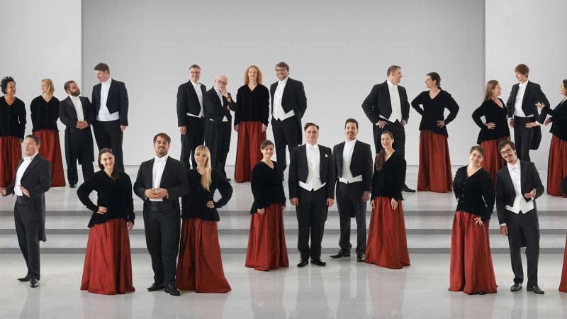 Der Kammerchor Stuttgart posiert in formeller Kleidung auf einer Bühne. Die Frauen tragen rote Röcke und schwarze Oberteile, die Männer Frack., © Jens Meisert