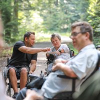Eine Gruppe von Menschen in Rollstühlen unterhält sich auf einem Waldweg. Die Umgebung ist grün und sonnig., © NPSFW