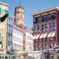 Bunte Gebäude und Marktstände auf einem Marktplatz, mit einem Kirchturm im Hintergrund unter blauem Himmel., © SMG, trickytine Bunte Gebäude und Marktstände auf einem Marktplatz, mit einem Kirchturm im Hintergrund unter blauem Himmel., © SMG, trickytine