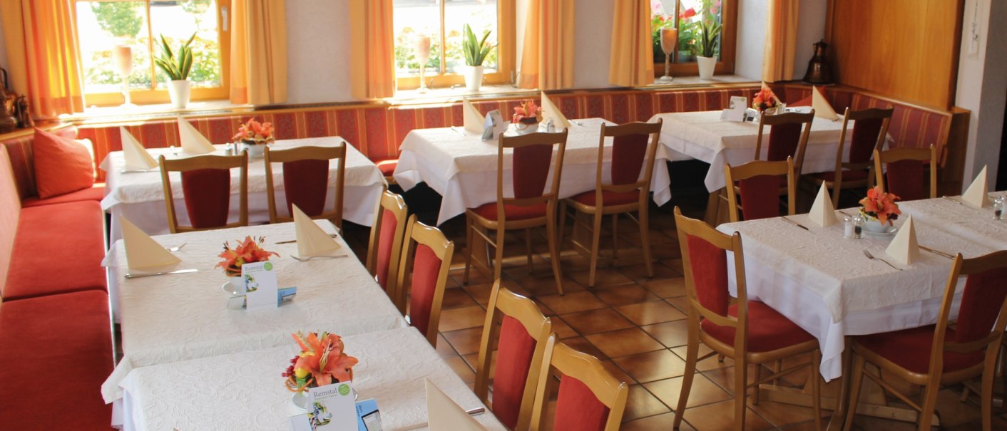 Gemütlicher Gastraum mit gedeckten Tischen, roten Stühlen und orangenen Vorhängen. Pflanzen und Dekorationen verleihen eine einladende Atmosphäre., © Gasthof zur Rose