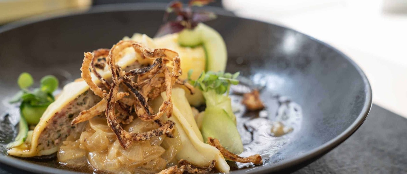 An elegant dish featuring stuffed pasta, crispy onions, and fresh herbs, served on a black plate., &copy; Flairhotel Vier Jahreszeiten