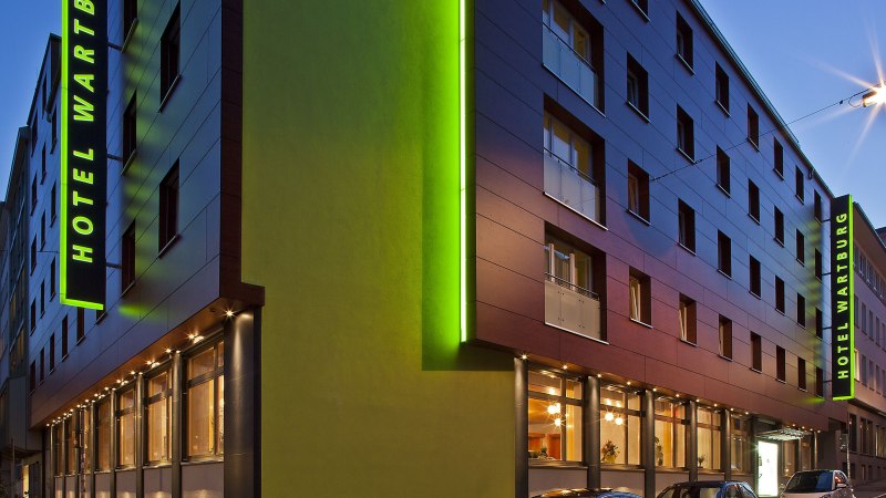 Moderne Hotelau&szlig;enansicht bei D&auml;mmerung, mit gr&uuml;ner Beleuchtung und parkenden Autos. Der Schriftzug 'Hotel Wartburg' ist sichtbar., &copy; VCH Hotel Wartburg