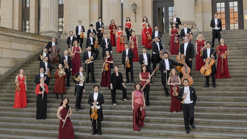 Orchester in formeller Kleidung posiert auf Treppe vor klassischem Geb&auml;ude. Musiker halten Instrumente, tragen rote und schwarze Outfits., &copy; Klassische Philharmonie Bonn e.V.