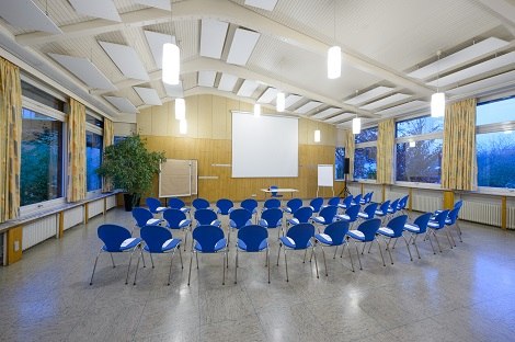 Heller Tagungsraum mit blauen Stühlen in Reihen, Projektorbildschirm, Flipchart und großen Fenstern mit Vorhängen., © Hotel am Schlossberg Herrenberg