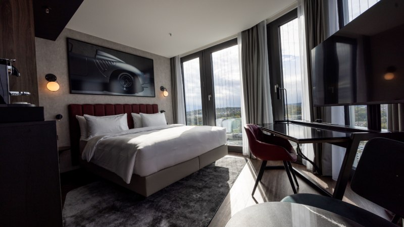 Modernes Eckzimmer mit gro&szlig;em Bett, Schreibtisch, Panoramafenstern und stilvollem Dekor. Ein Bild eines Autos h&auml;ngt an der Wand., &copy; Radisson Hotel Group