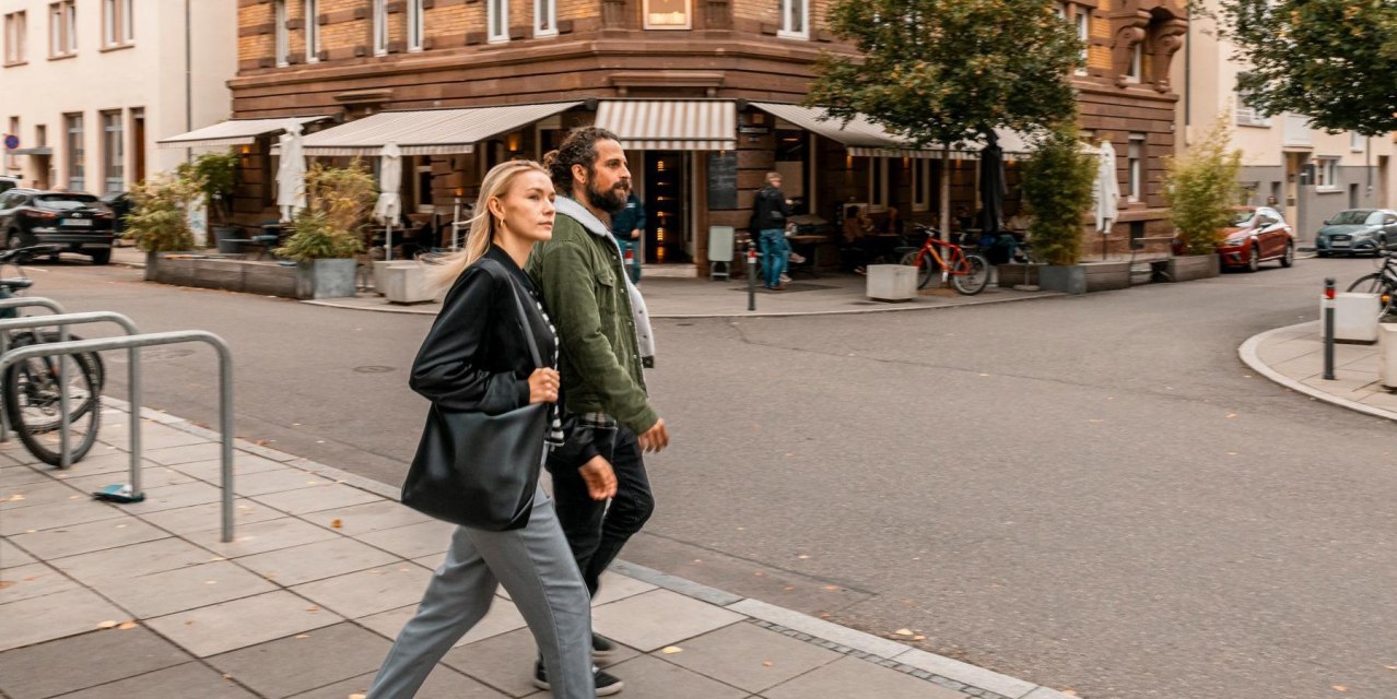 Ein Mann und eine Frau überqueren eine Straße in einer städtischen Umgebung. Im Hintergrund ist ein Café mit Außensitzplätzen zu sehen., © SMG, Sarah Schmid Ein Mann und eine Frau überqueren eine Straße in einer städtischen Umgebung. Im Hintergrund ist ein Café mit Außensitzplätzen zu sehen., © SMG, Sarah Schmid