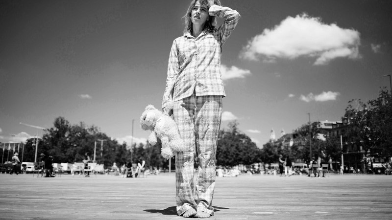 Eine Frau in kariertem Pyjama steht auf einem gro&szlig;en Platz und h&auml;lt ein Pl&uuml;schtier. Der Himmel ist klar mit einigen Wolken, und im Hintergrund sind B&auml;ume und Geb&auml;ude zu sehen., &copy; Rosenau Kultur e.V.