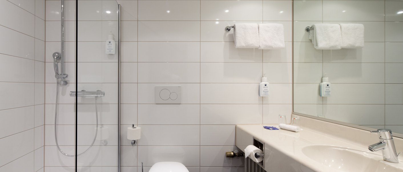 Modernes Badezimmer mit weiß gefliesten Wänden, Glasdusche, Toilette, Waschbecken und Spiegel. Handtücher und Seifenspender sind vorhanden., © Erikson Hotel Modernes Badezimmer mit weiß gefliesten Wänden, Glasdusche, Toilette, Waschbecken und Spiegel. Handtücher und Seifenspender sind vorhanden., © Erikson Hotel