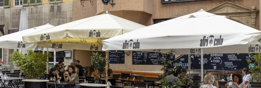 Outdoor area of a café called Platzhirsch with parasols and guests. People sit at tables and enjoy drinks., © SMG, Sarah Schmid