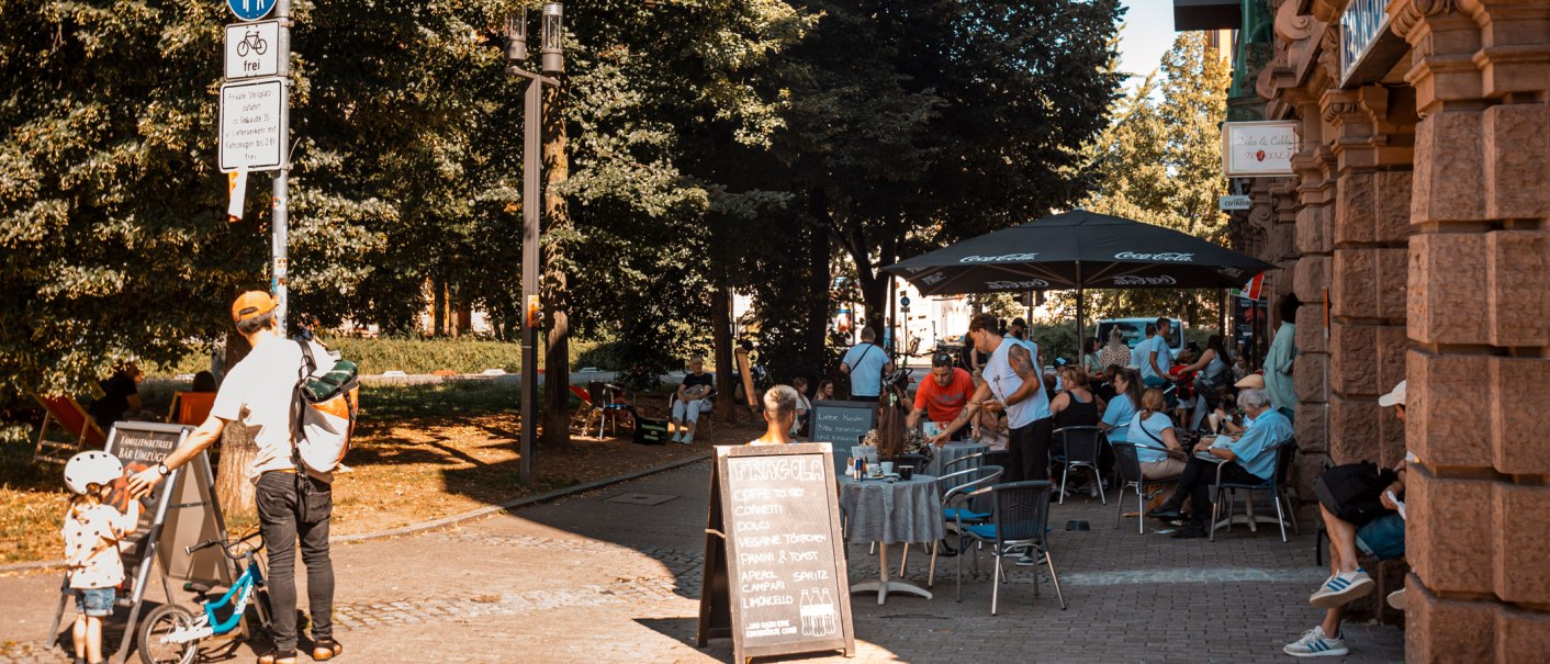 Straßencafé am Bismarckplatz mit Menschen, die im Freien sitzen. Ein Mann mit Kind und Fahrrad steht neben einem Schild. Bäume spenden Schatten., © SMG, Sarah Schmid