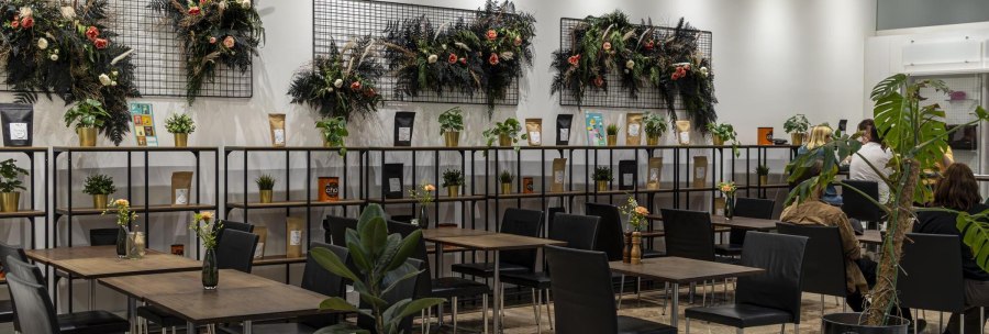 Modern café with plants and decorative wall arrangements. Black chairs and wooden tables, shelves with products and plants., © SMG Stuttgart Marketing GmbH - Sarah Schmid