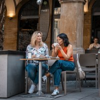 Zwei Frauen sitzen in einem Straßencafé, trinken Kaffee und unterhalten sich. Im Hintergrund sind weitere Gäste und ein Café-Eingang zu sehen., © Stuttgart Marketing GmbH - Sarah Schmid Zwei Frauen sitzen in einem Straßencafé, trinken Kaffee und unterhalten sich. Im Hintergrund sind weitere Gäste und ein Café-Eingang zu sehen., © Stuttgart Marketing GmbH - Sarah Schmid