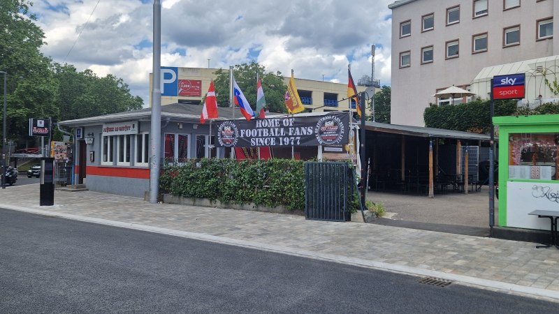 Bar mit Fahnen und Schild "Home of Football Fans Since 1977". Gebäude mit roten und grauen Akzenten, umgeben von Bäumen und einem Parkplatz., © Stuttgart-Marketing GmbH Bar mit Fahnen und Schild "Home of Football Fans Since 1977". Gebäude mit roten und grauen Akzenten, umgeben von Bäumen und einem Parkplatz., © Stuttgart-Marketing GmbH