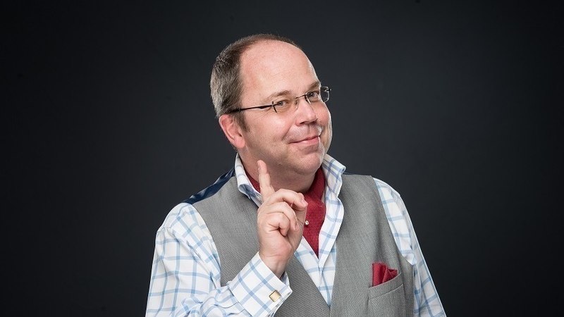 Ein Mann in kariertem Hemd und grauer Weste hebt l&auml;chelnd den Zeigefinger vor einem dunklen Hintergrund., &copy; Erlebnis Schmaus