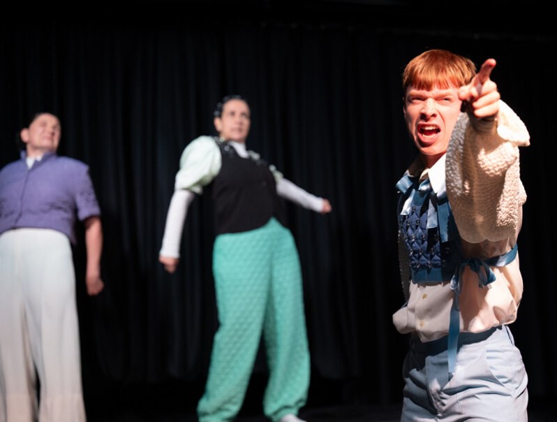 Drei Personen auf einer Bühne, eine im Vordergrund zeigt energisch nach vorne. Die anderen stehen im Hintergrund vor einem schwarzen Vorhang., © Württembergische Staatstheater Stuttgart Drei Personen auf einer Bühne, eine im Vordergrund zeigt energisch nach vorne. Die anderen stehen im Hintergrund vor einem schwarzen Vorhang., © Württembergische Staatstheater Stuttgart