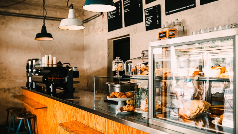 Ein gemütliches Café mit Holztresen, Kaffeemaschine und Gebäck in einer Glasvitrine. Hängelampen beleuchten den Raum., © Melanie Mearz Fotografie