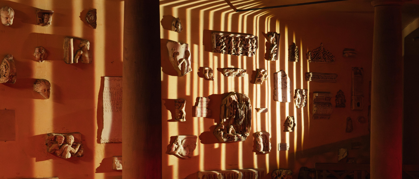 Antike Reliefs und Skulpturen an einer orangefarbenen Wand, beleuchtet von Streifenlicht, das durch eine Säule fällt. Die Szene wirkt warm und atmosphärisch., © Lichtgestalten