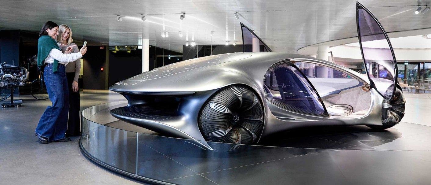Two people pose for a selfie in front of a futuristic car with open wing doors in a modern showroom., © Mercedes-Benz AG