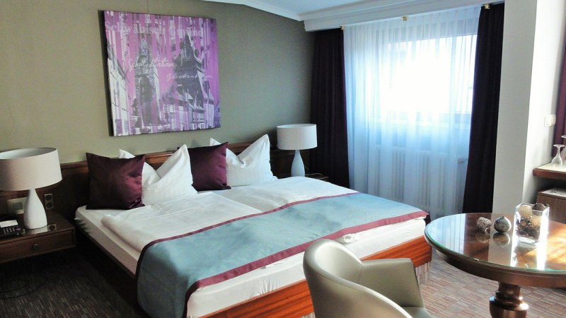 Komfortables Doppelzimmer mit großem Bett, zwei Nachttischen, Tisch und Stühlen. Helle Vorhänge und moderne Dekoration an der Wand., © Hotel Pelikan