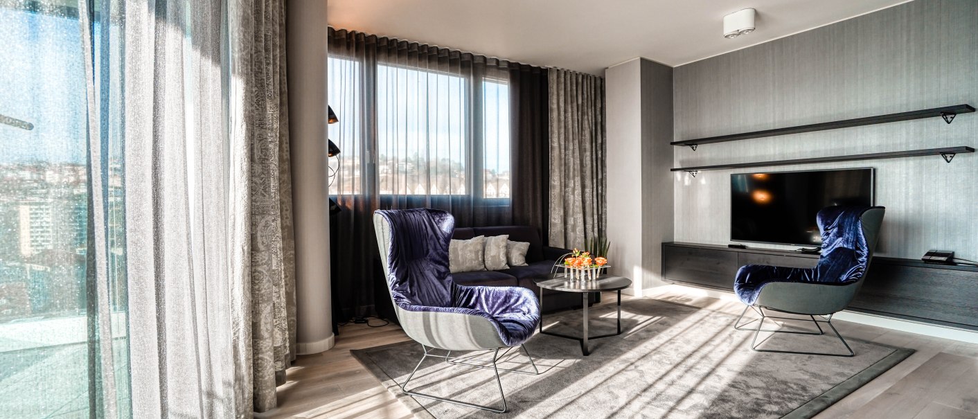 Moderne Suite mit großen Fenstern, blauen Sesseln, Flachbildfernseher und Couchtisch auf Teppich., © CLOUD N°7 APARTMENTS GmbH Moderne Suite mit großen Fenstern, blauen Sesseln, Flachbildfernseher und Couchtisch auf Teppich., © CLOUD N°7 APARTMENTS GmbH