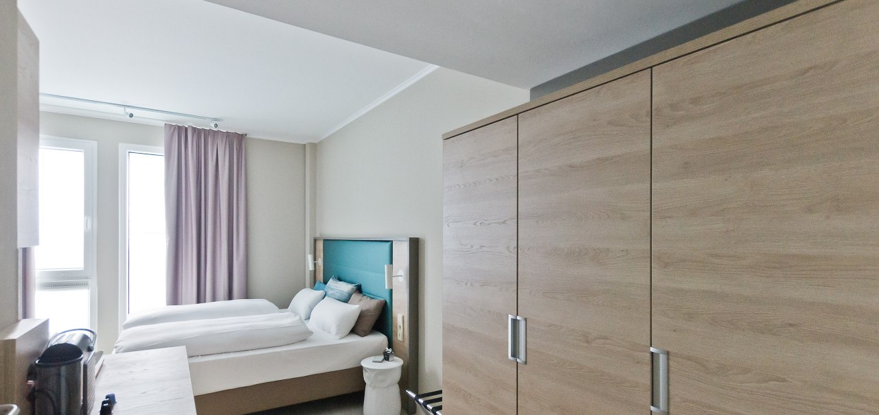 Ein modernes Hotelzimmer mit einem Doppelbett, einem gro&szlig;en Kleiderschrank und zwei Fenstern mit Vorh&auml;ngen. Ein Tisch mit Wasserkocher ist sichtbar., &copy; Arthotel Ana / elaya