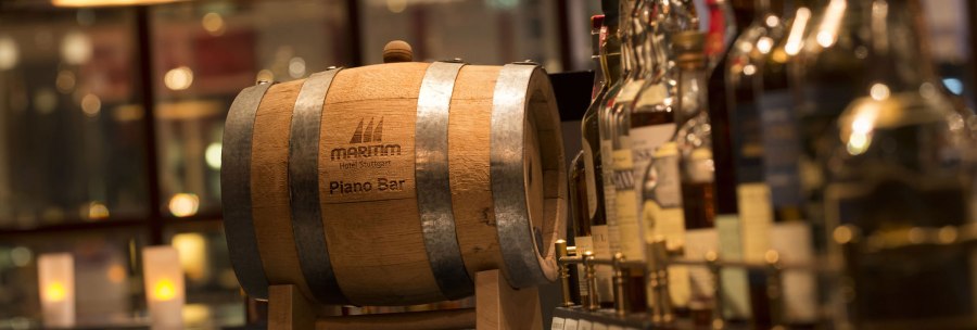 Ein Holzfass mit der Aufschrift 'Maritim Hotel Stuttgart Piano Bar' steht auf einer Bar, umgeben von verschiedenen Flaschen und Kerzenlicht., &copy; Maritim Hotels