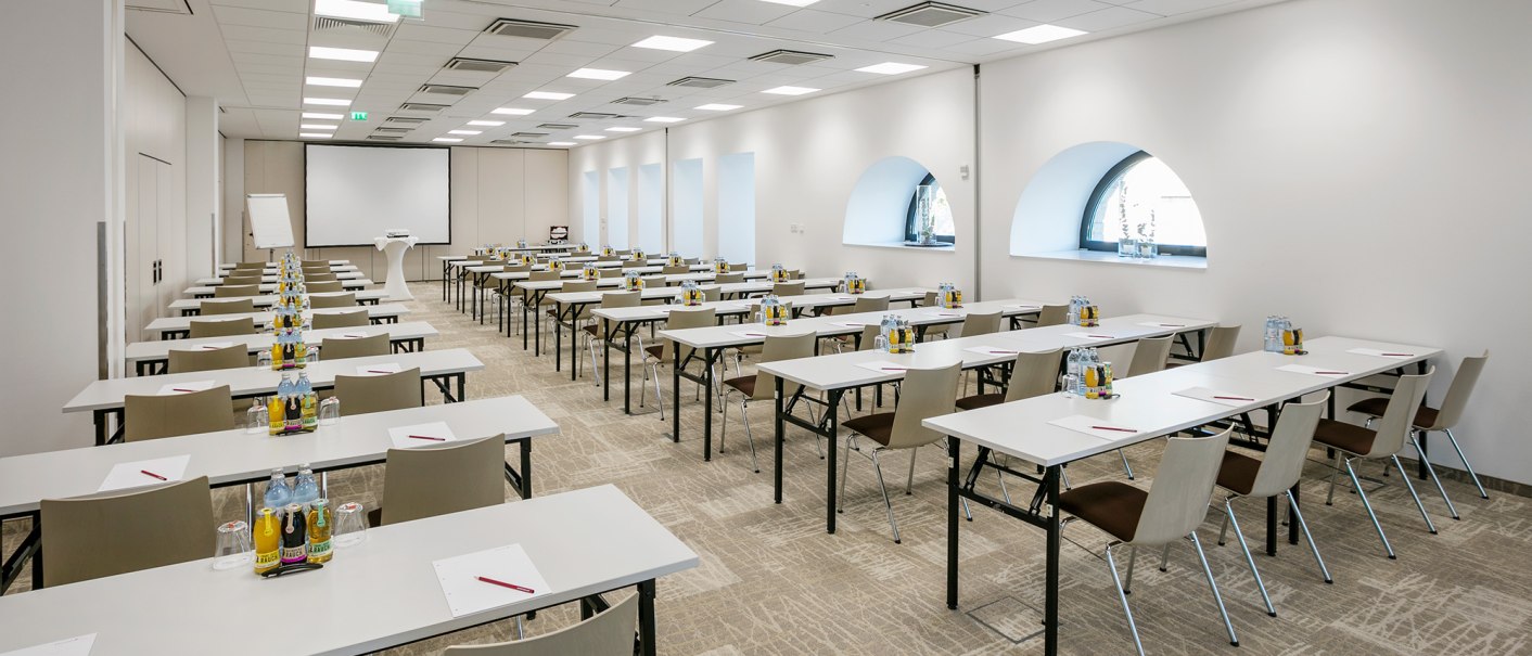 Moderner Tagungsraum mit weißen Tischen und Stühlen, ausgestattet mit Getränken und Schreibmaterialien. Ein Projektor und eine Leinwand sind vorhanden., © ARCOTEL Hotels