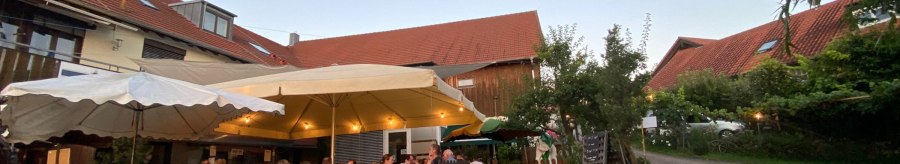 Menschen sitzen unter großen Sonnenschirmen in einem gemütlichen Hof mit roten Dächern und Bäumen., © Weinhof Zaiß